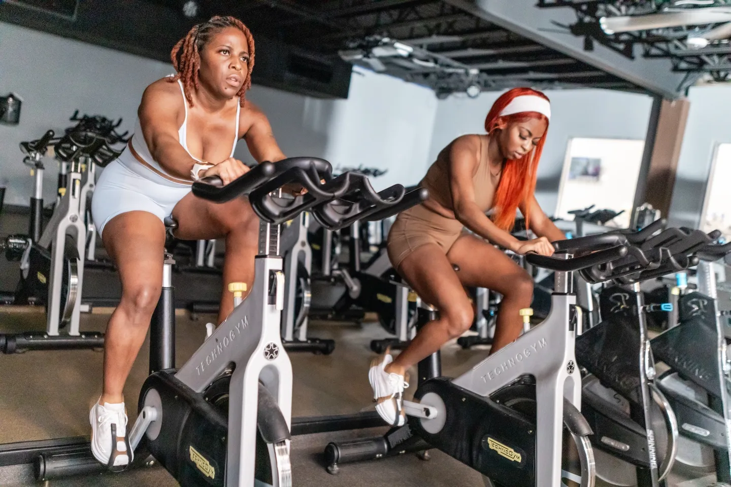 Two women are riding stationary bikes in a gym.